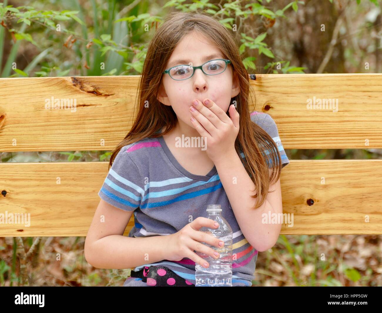 Young girl on wooden bench with hand to mouth, holding a clear water bottle.  Gainesville, Florida, USA. Stock Photo