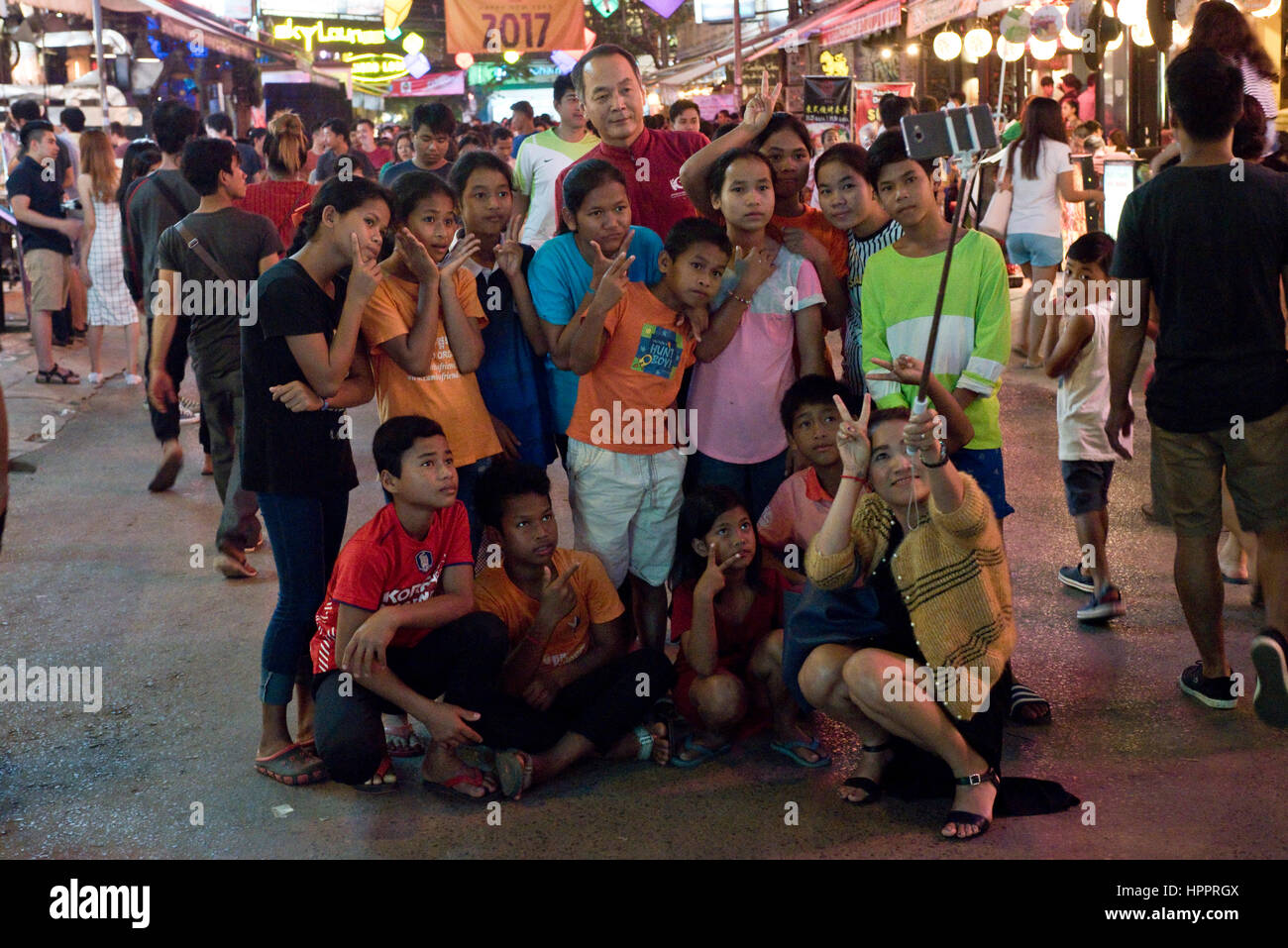 A group family of Asian tourists on Pub Street, Siem Reap, taking a self portrait with a selfie stick and mobile phone on a busy night street scene. Stock Photo