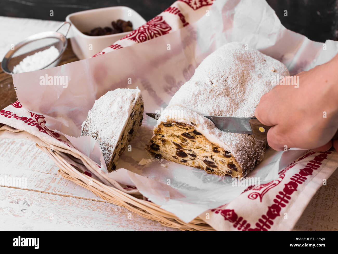 Woman's hand cutting off slice of powdered Christmas stollen on parchment paper, wicker basket, kitchen towel, ingredients, rustic kitchen interior Stock Photo