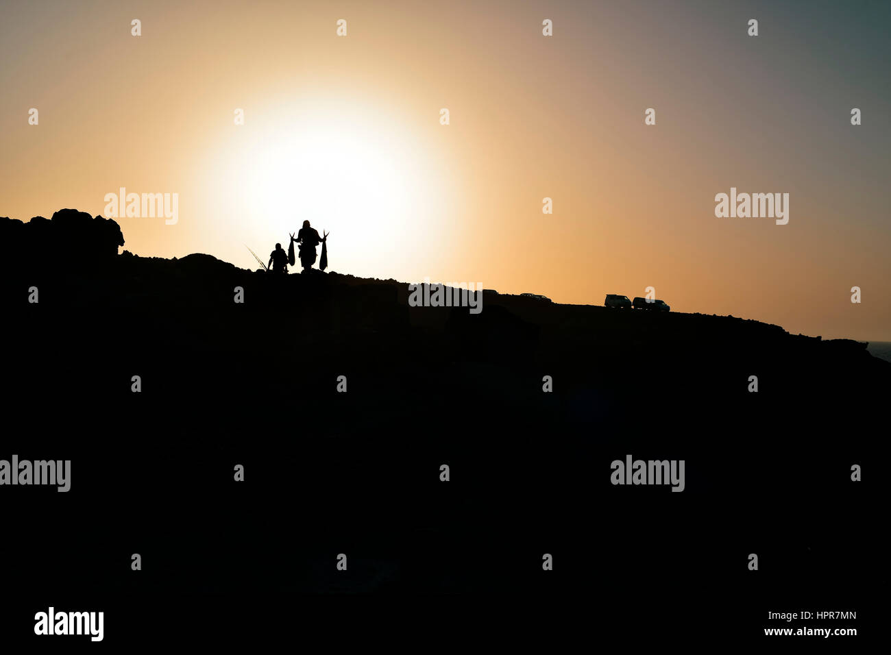 the silhouette of two fishermen carrying fishes and fishing rods at the top of a cliff at dusk, against a colorful yellow sky Stock Photo