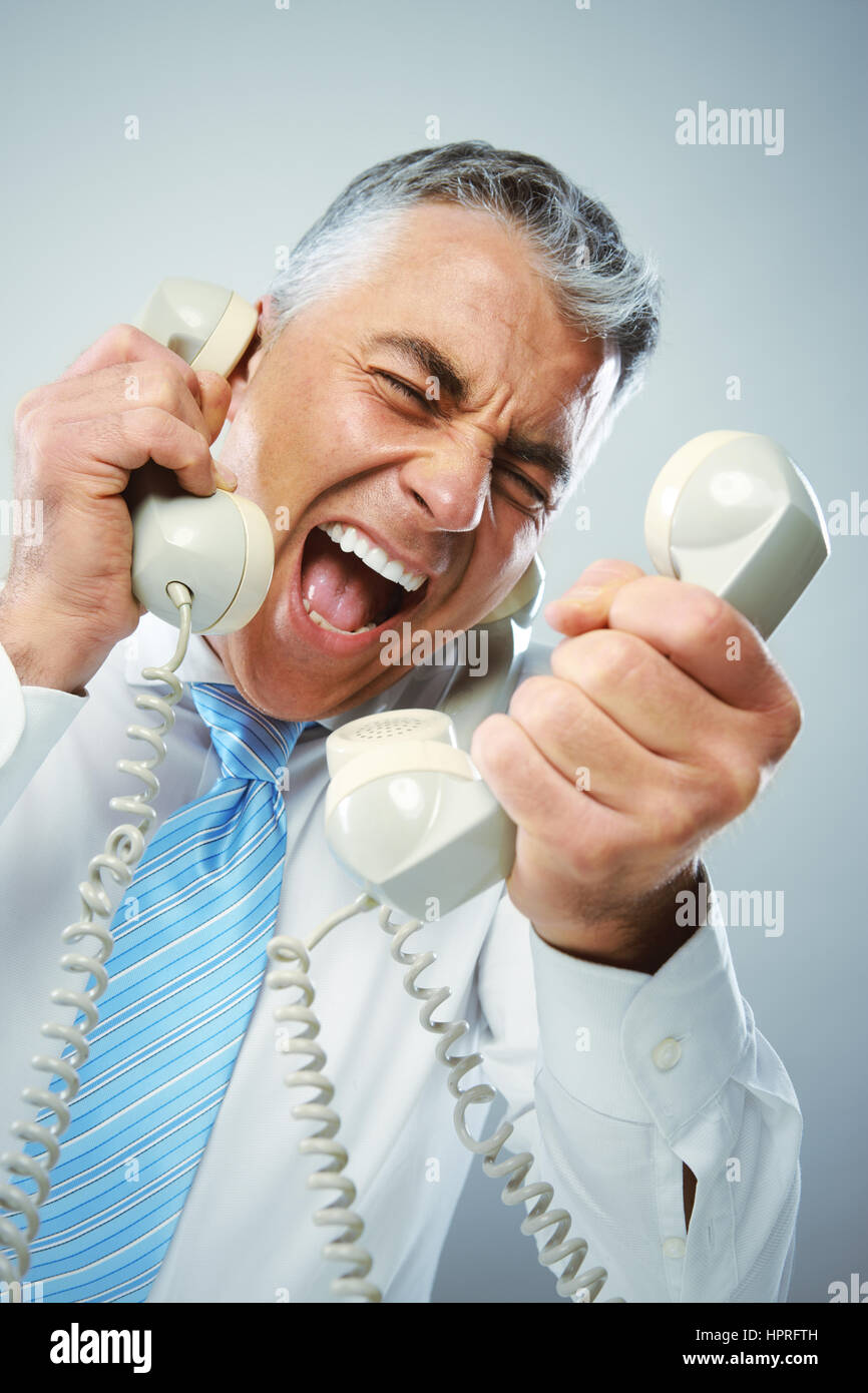 A stressed businessman yells loudly into the three handsets that he is holding. Stock Photo