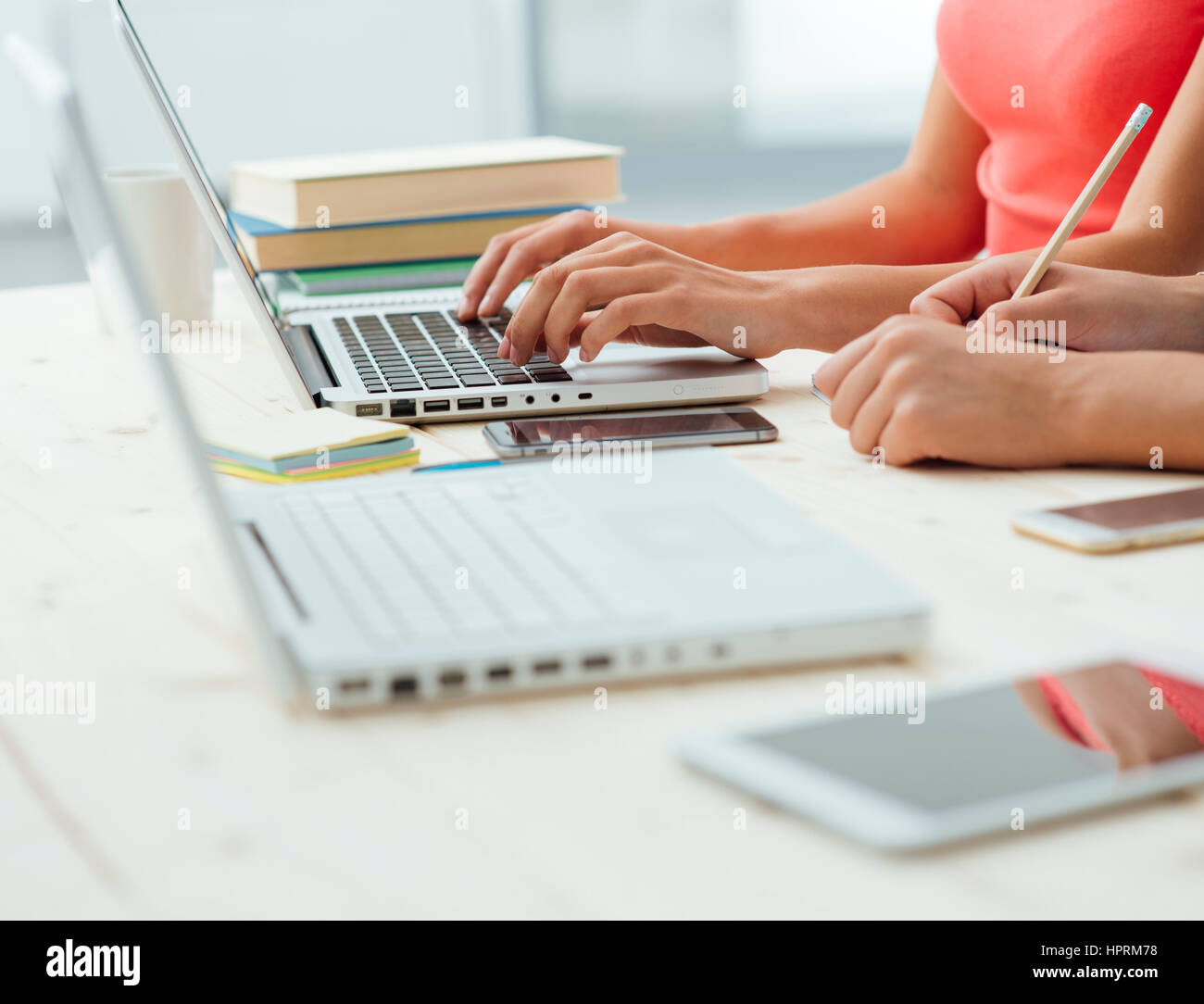 Teen girls studying at desk and doing homeworks, one is using a laptop and the other one is writing on a notebook, education concept, unrecognizable p Stock Photo