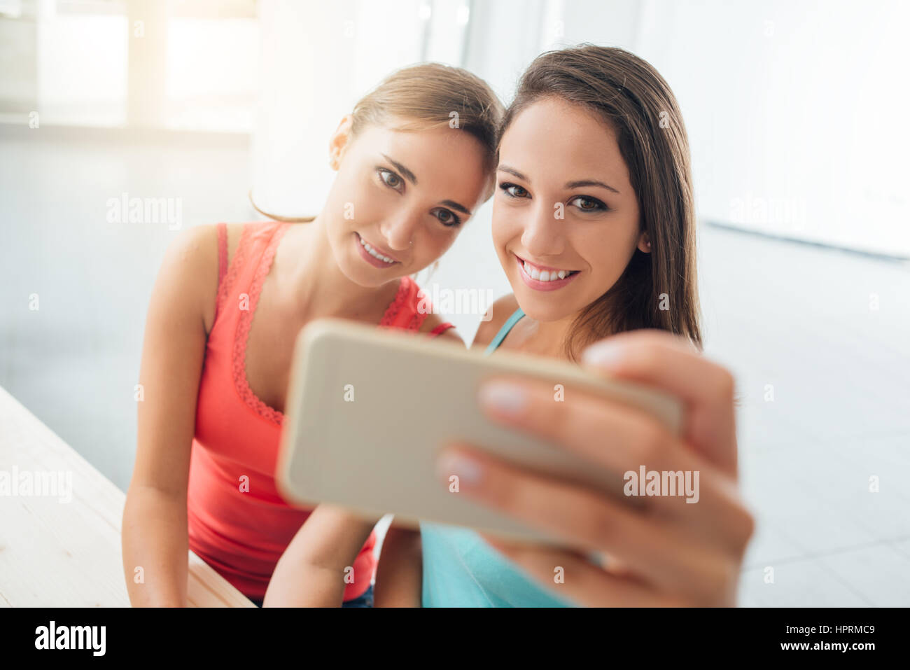 Cute girls sitting at desk and taking self portraits using a smart phone, they are smiling at camera and posing Stock Photo
