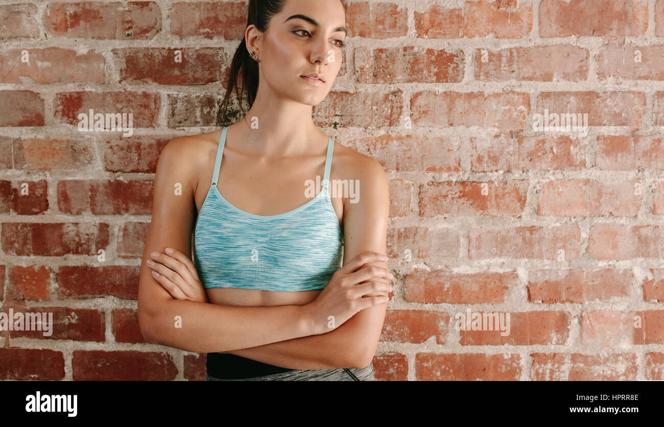Portrait of fit young woman in sports bra standing against brick wall with her arms crossed. Caucasian female fitness model in gym looking away. Stock Photo