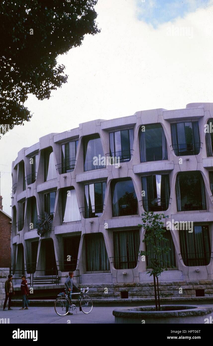 People stand outside of a circular building with windows inset into the concrete facade that has a design that alternates both horizontally and vertically, 1965. Stock Photo