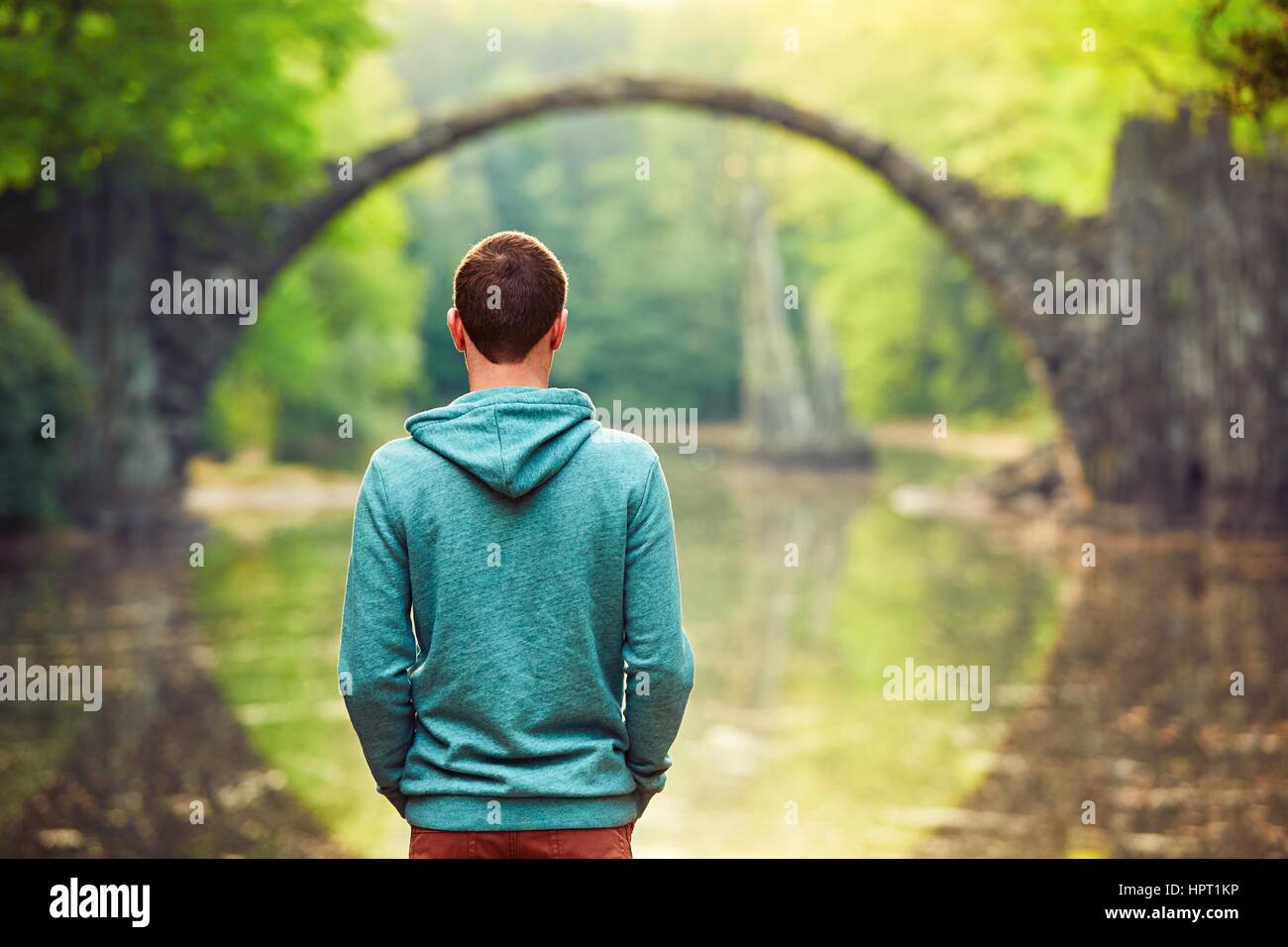 Pensive young man on the pond bank. - Amazing place in Germany. Rakotzbrucke also known as Devils Bridge in Kromlau. Stock Photo