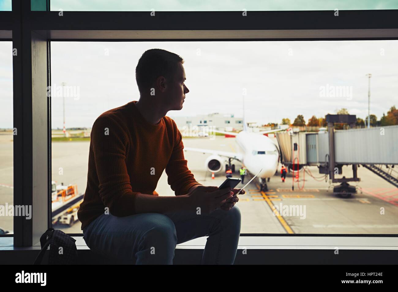 Silhouette of the young man with mobile phone and boarding pass in hand waiting inside airport terminal. The ground crew prepares the plane for the ne Stock Photo