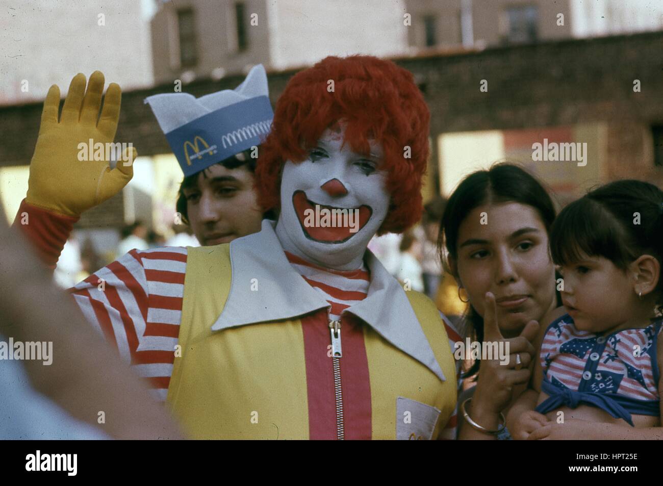 A performer dressed as Ronald McDonald waves at a camera while a Latina woman and child stand nearby and look on in the Bronx, New York City, New York, 1976. Stock Photo