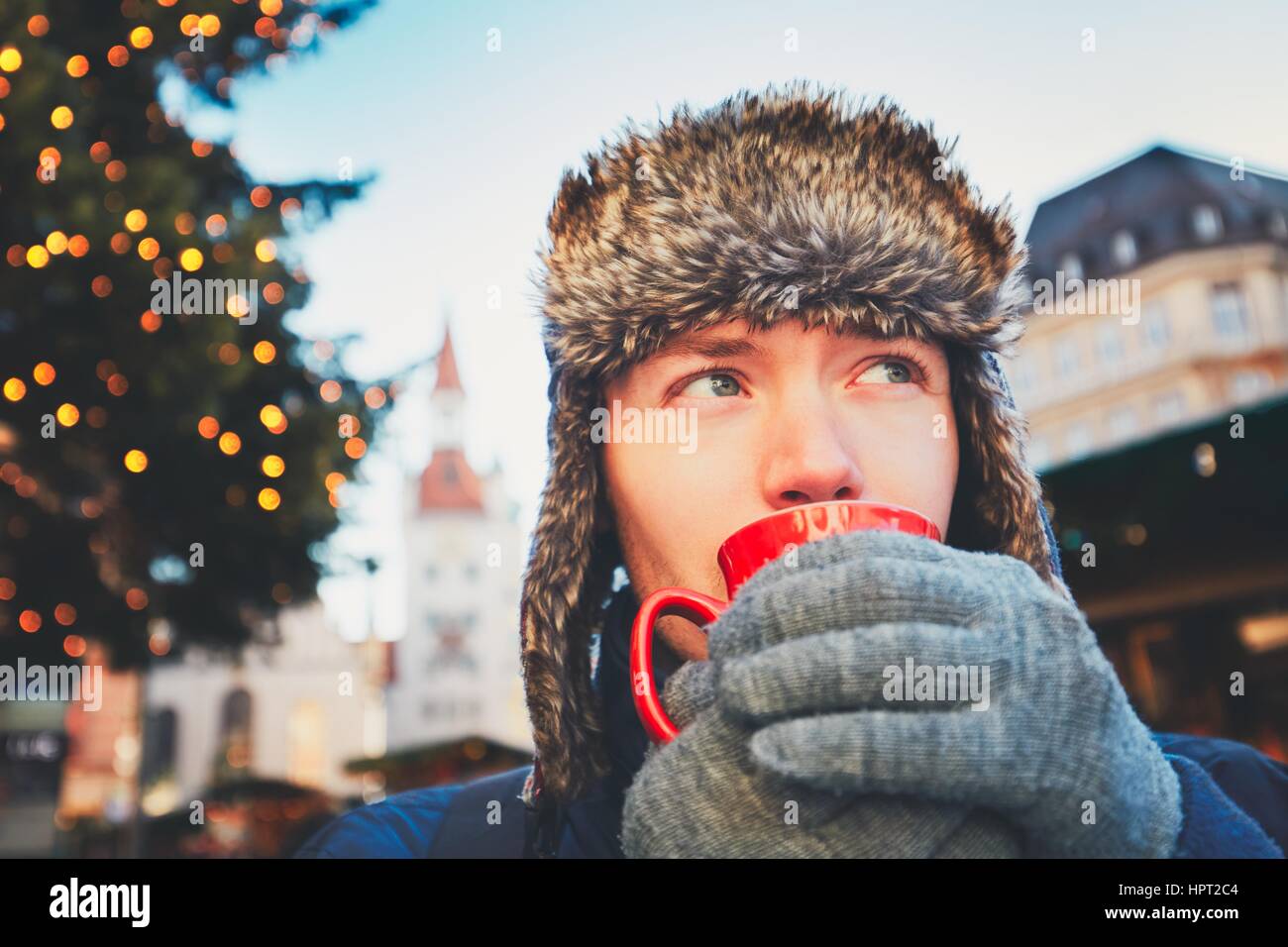 Young man drinking hot wine (or punch or tea or coffee) from red cup at the Christmas market - Munich, Germany Stock Photo