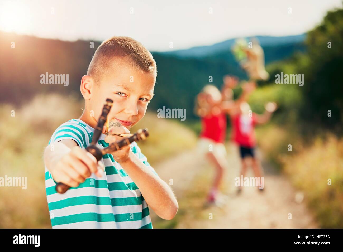 Little boy holding slingshot with stone. Boy is playing with his sisters in rural landscape. Stock Photo