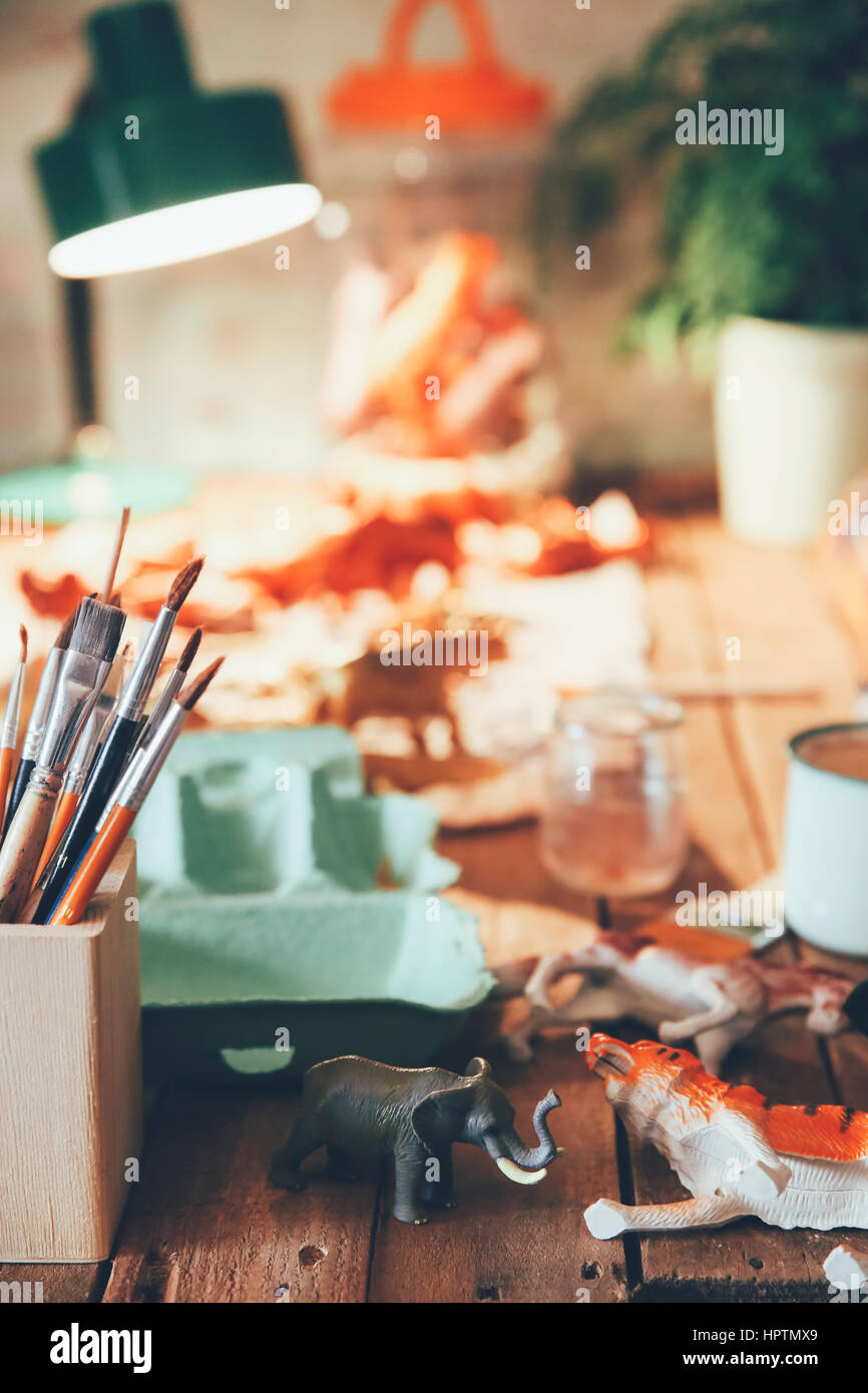 Table with pot of paintbrushes, plastic animals, lamp and plant in the background Stock Photo