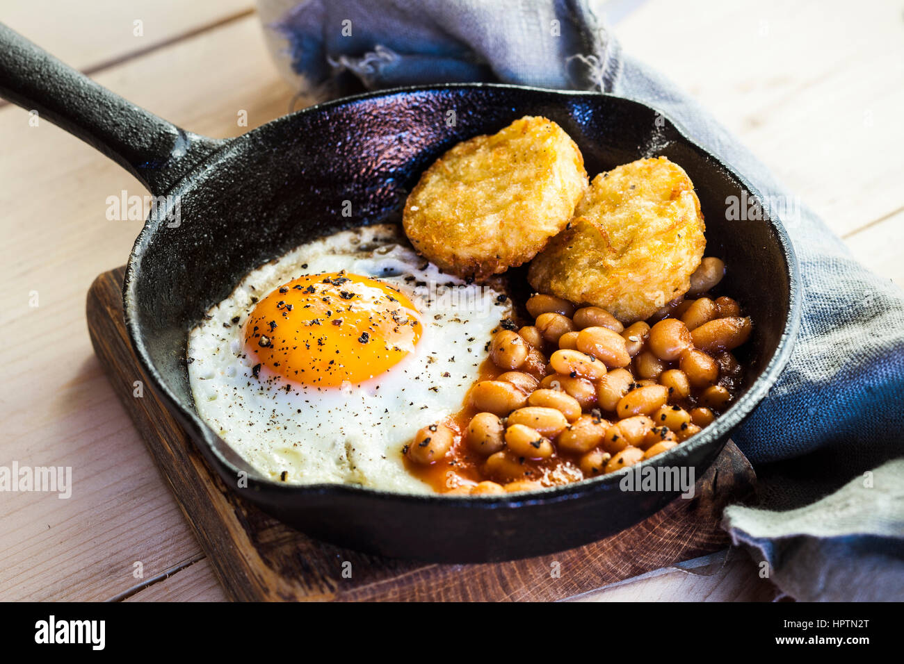 Fried egg, baked beans and hash browns in frying pan on wooden board