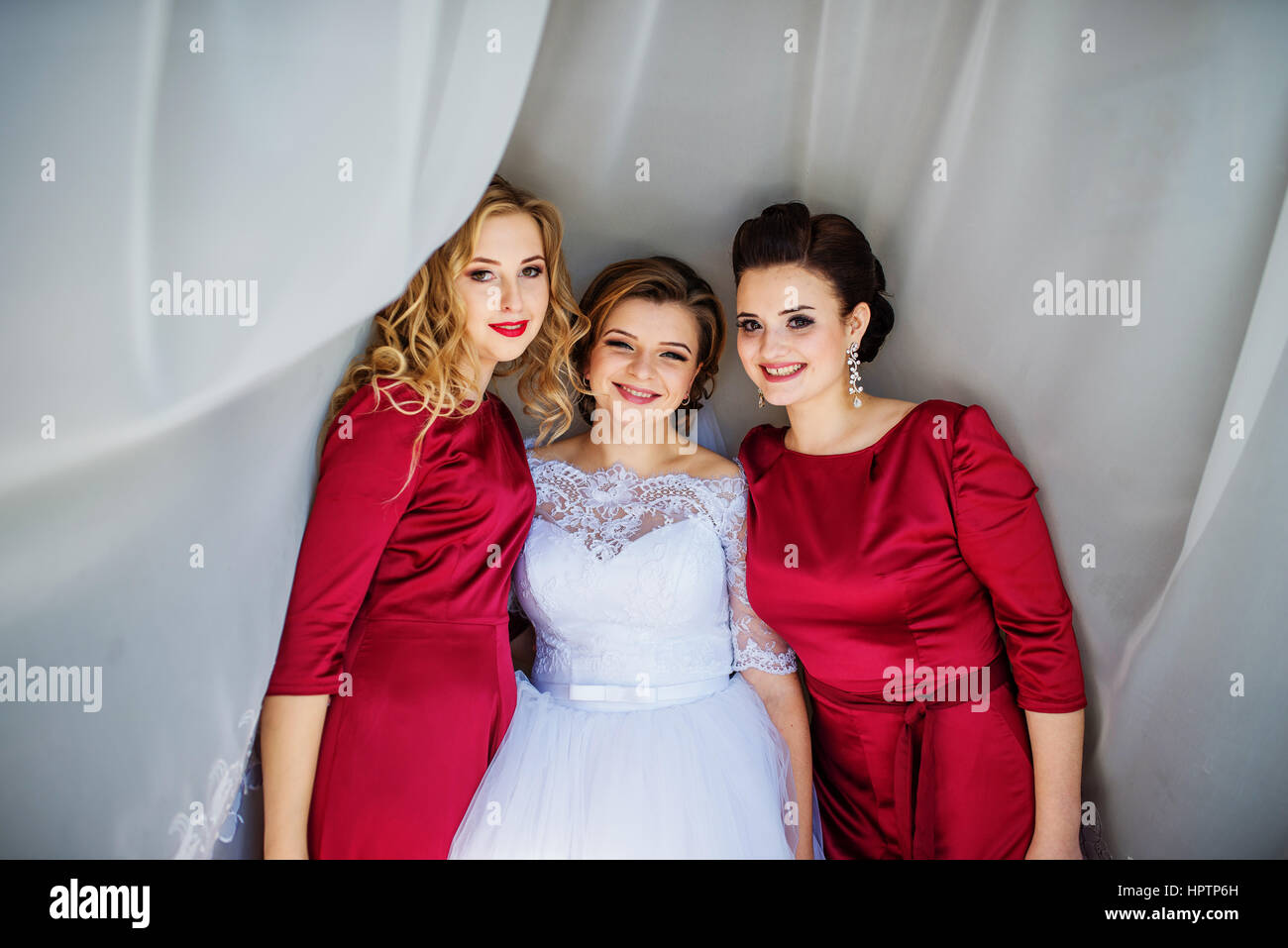 Cute blonde bride with bridesmaids posed on curtains at wedding morning day. Stock Photo