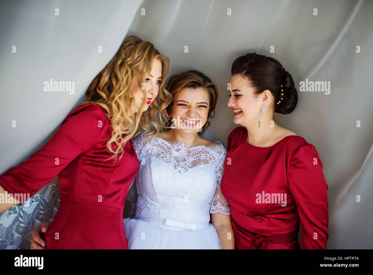 Cute blonde bride with bridesmaids posed on curtains at wedding morning day. Stock Photo