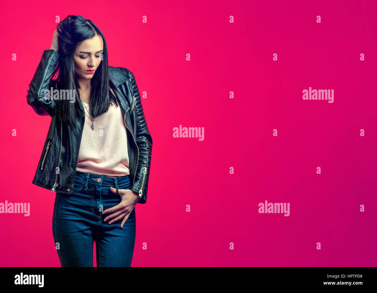 Brooding stylish girl with black hair in a leather jacket and blue jeans on a bright pink background. Stock Photo