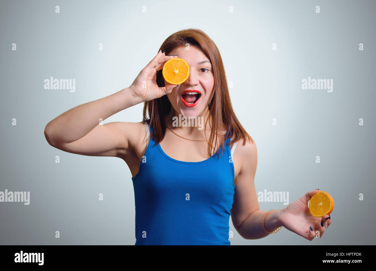 Cheerful young girl with a slice of orange around the eyes on the gray studio background. Stock Photo