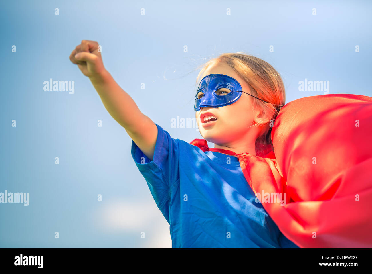 Funny little girl playing power super hero over blue sky background. Superhero concept. Stock Photo