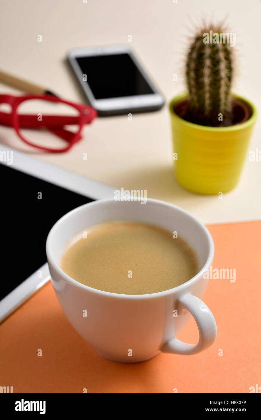 closeup of an office desk full of things, such as a cup with white coffee, a pen, a tablet, a smartphone, a cactus and a pair of red eyeglasses Stock Photo