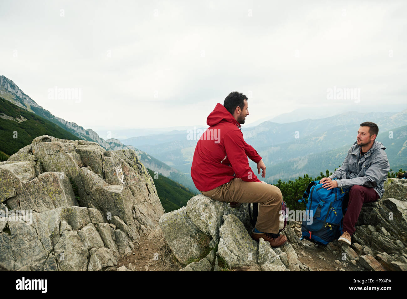 Two men in hiking gear sitting on a rock high in the mountains talking ...