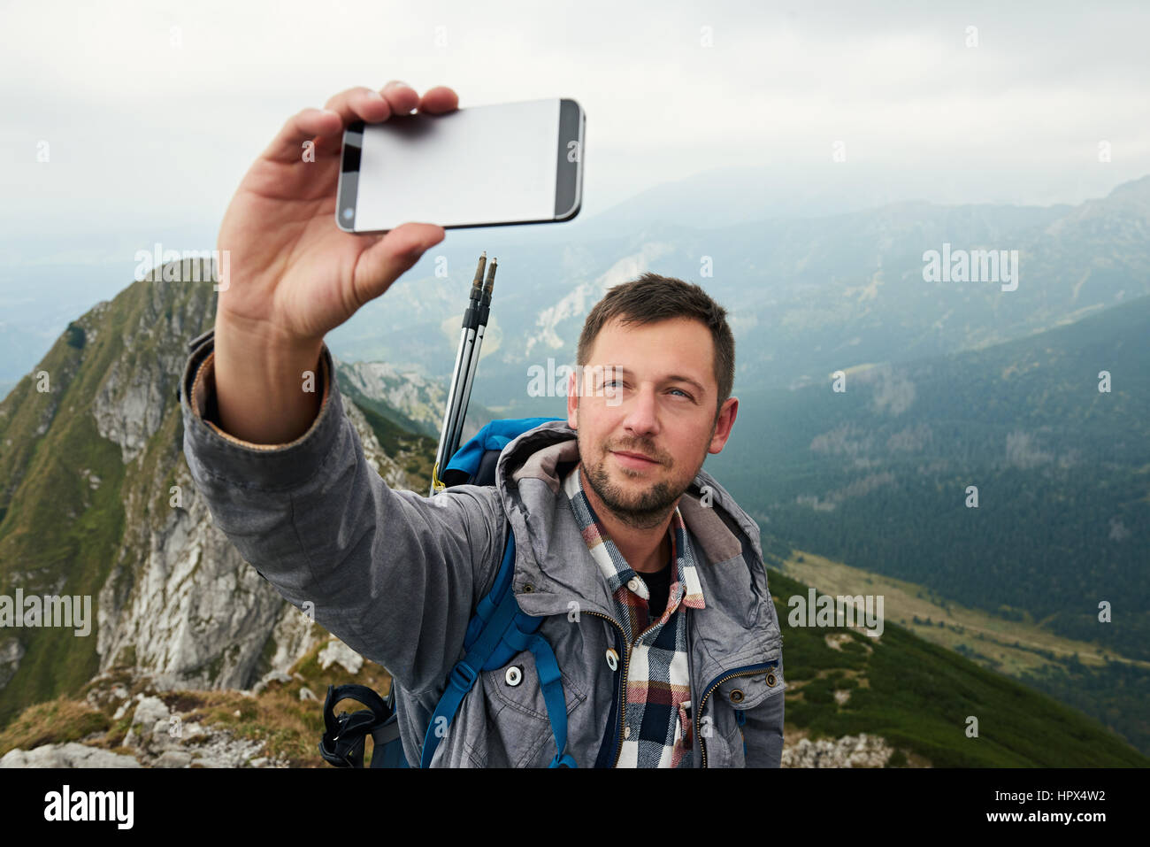 Man in hiking gear standing atop a mountain taking a selfie with a ...