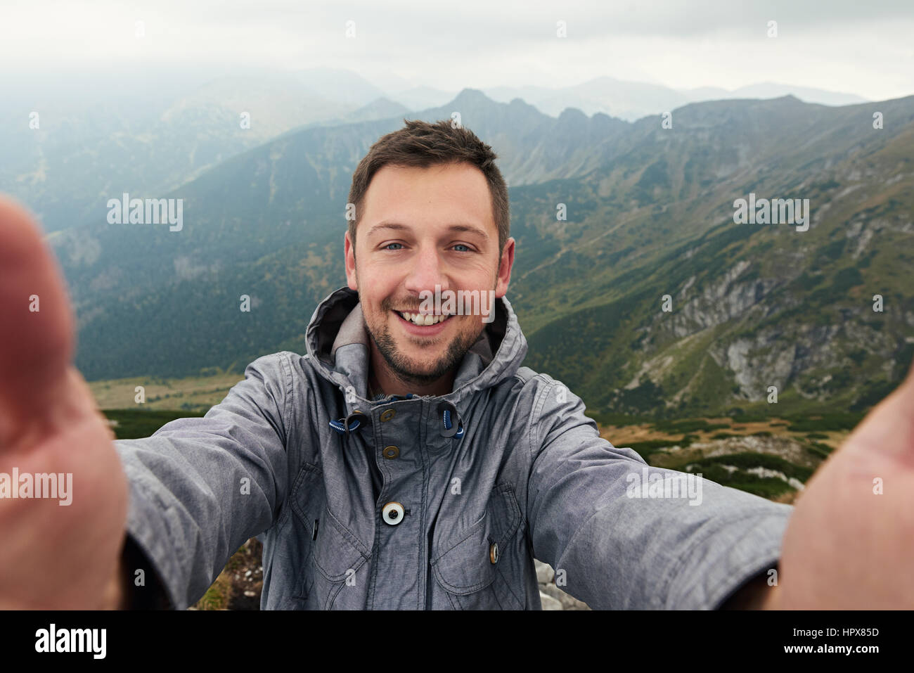 Smiling man in hiking gear standing atop a mountain taking a selfie ...
