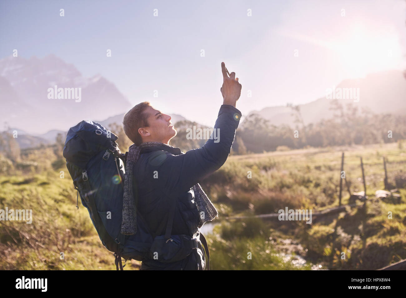 Young man with backpack hiking and using camera phone in sunny, remote field Stock Photo