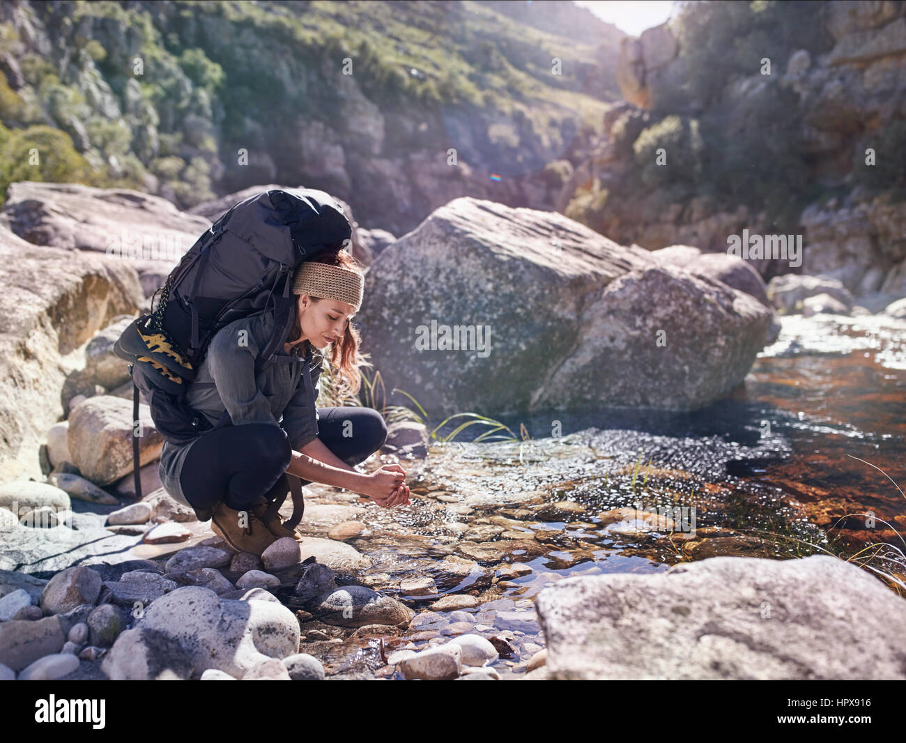 Young woman with backpack hiking, washing hands at sunny stream Stock Photo