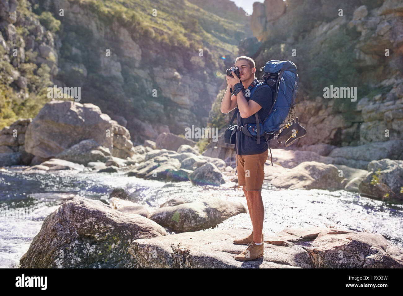 Young man with backpack hiking, photographing with camera on sunny rocks Stock Photo