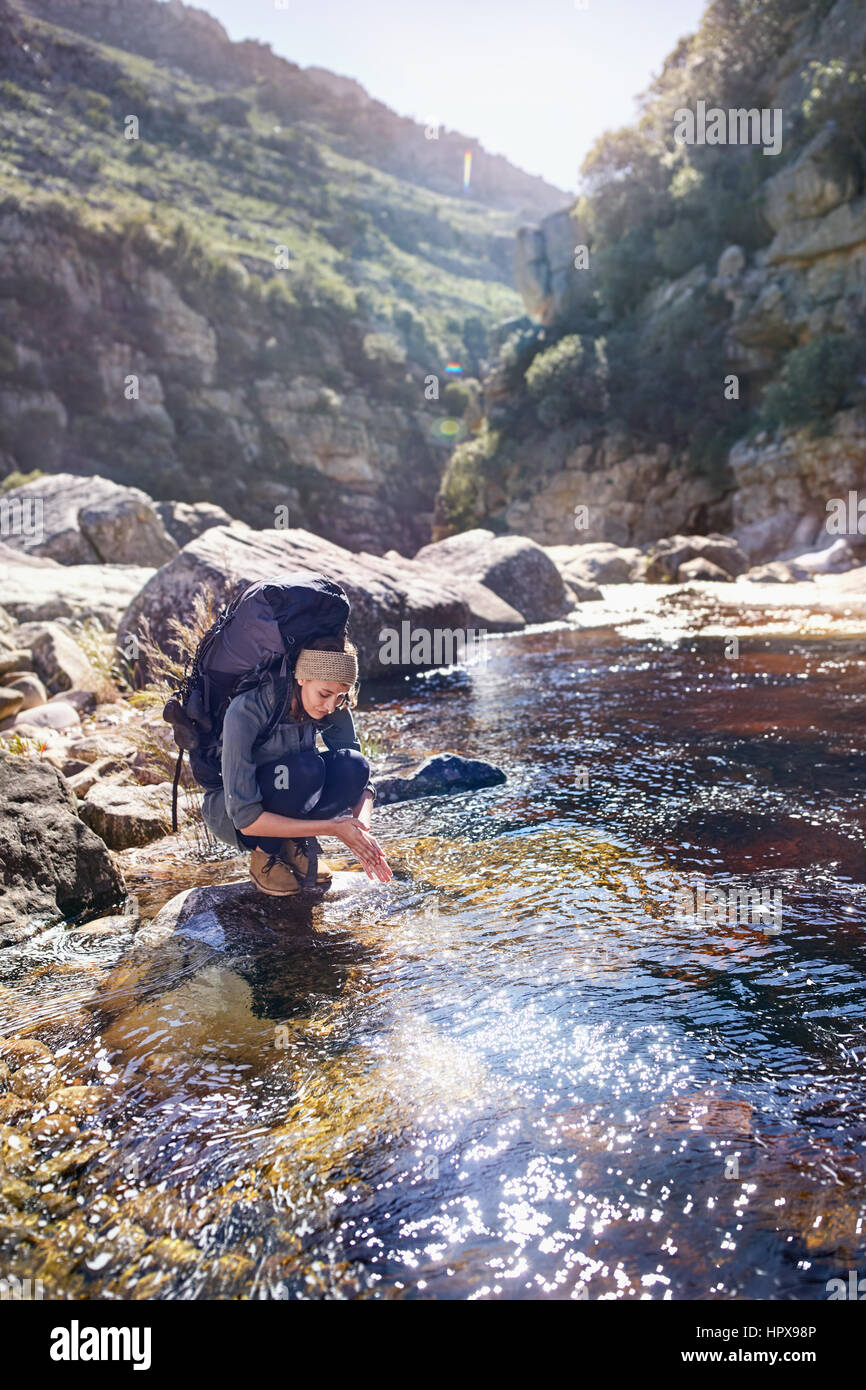 Young woman with backpack hiking, washing hands at sunny stream Stock Photo