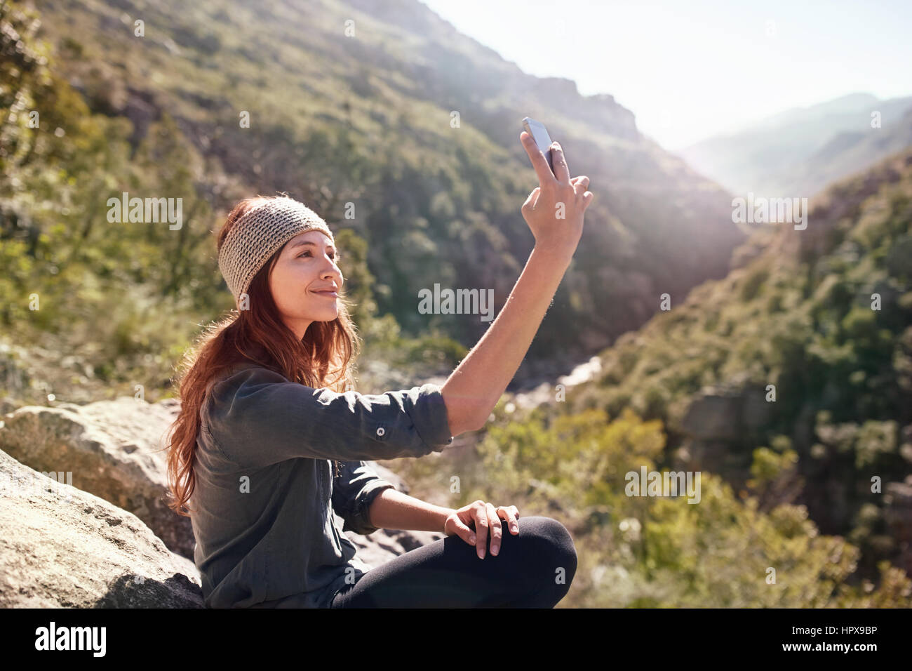 Young woman taking selfie with camera phone on sunny, remote rock Stock Photo