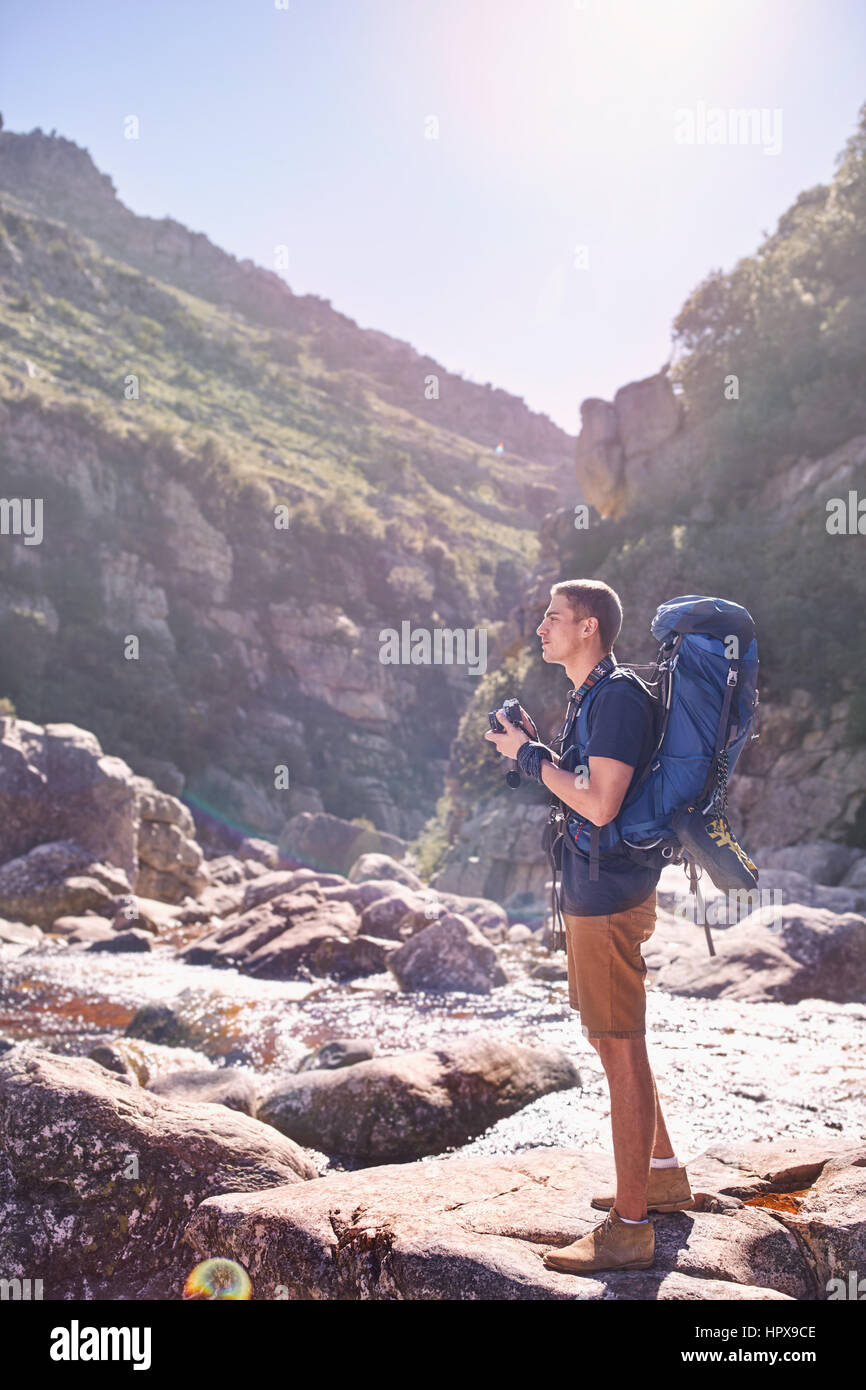 Young man with backpack hiking and photographing sunny, craggy cliffs with camera Stock Photo