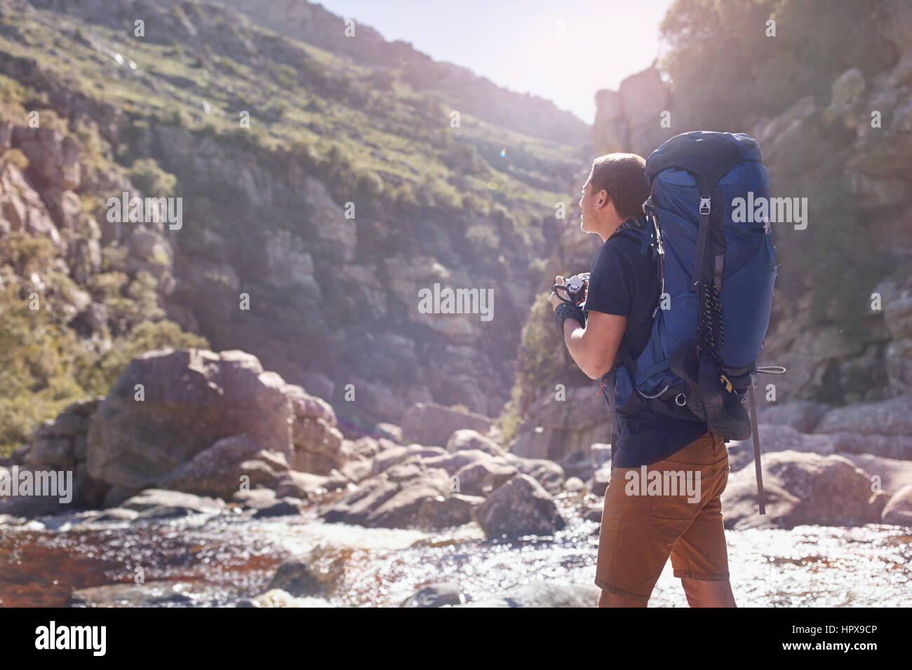 Young man with backpack hiking photographing with camera below sunny, remote cliffs Stock Photo