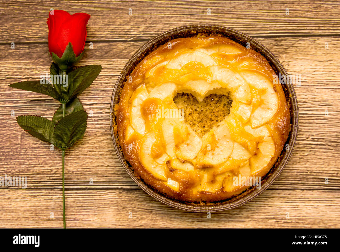 Apple pie with a hollow in the shape of a heart on a wooden table in which there is a red rose Stock Photo