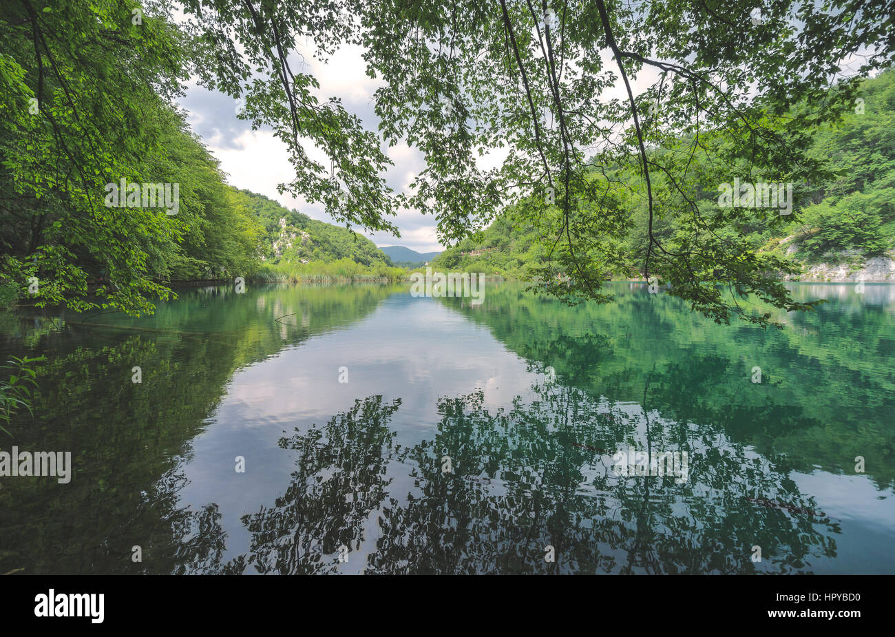 Trees, sky and mountains reflect in the crystal clear lakes of Plitvice Lakes National Park, as tourists walking along the boardwalk in the distance. Stock Photo