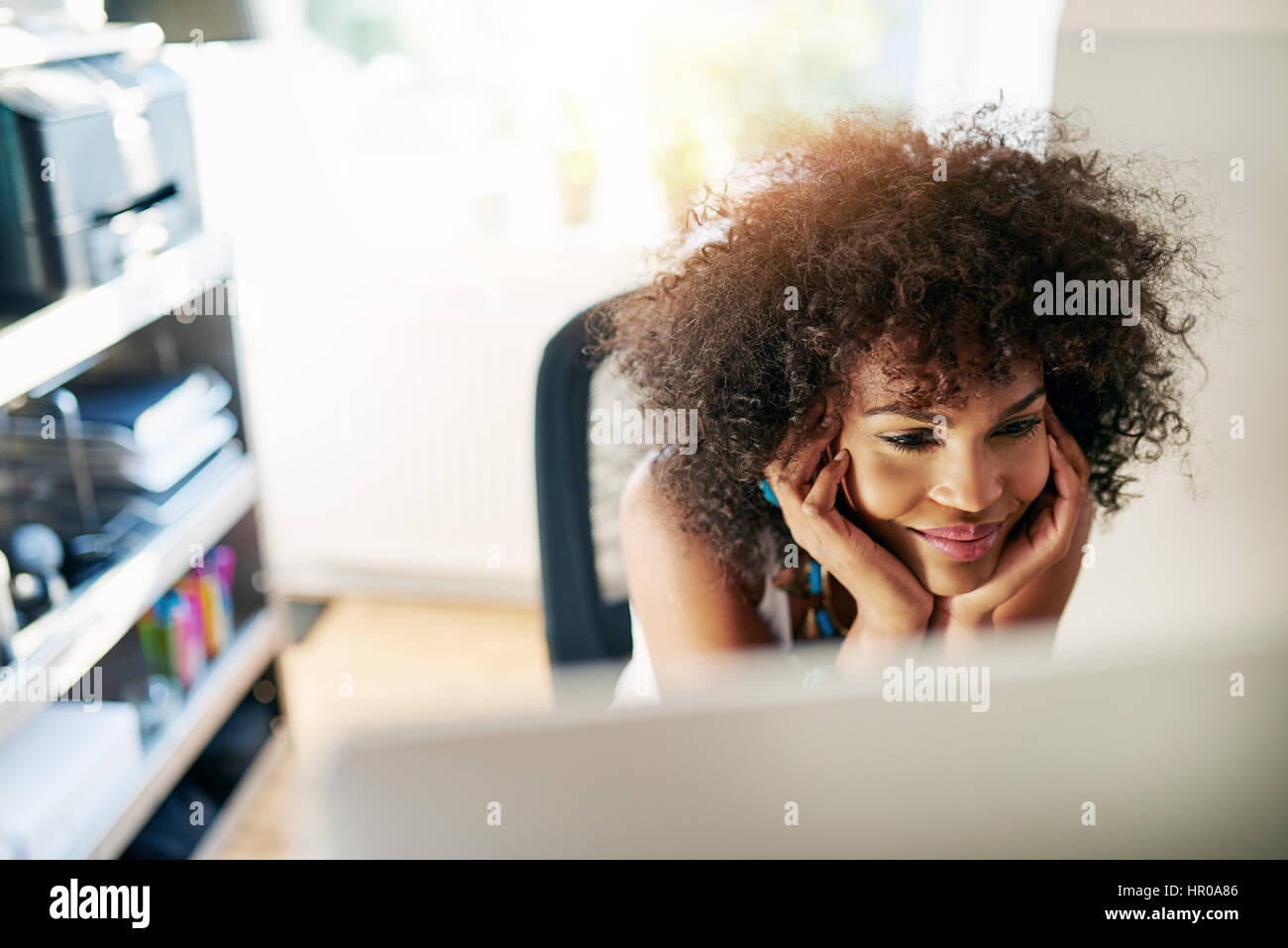 Young pretty afro-american woman looking at desk-top and smiling on blurred inside background. Stock Photo