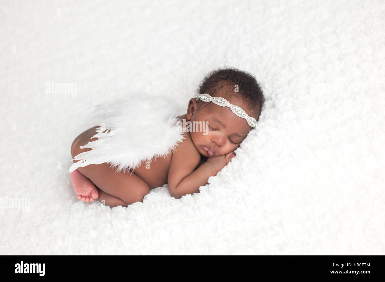 Portrait of a one month old, sleeping, newborn, baby girl. She is wearing a rhinestone headband, feather angel wings, and sleeping on a white blanket. Stock Photo