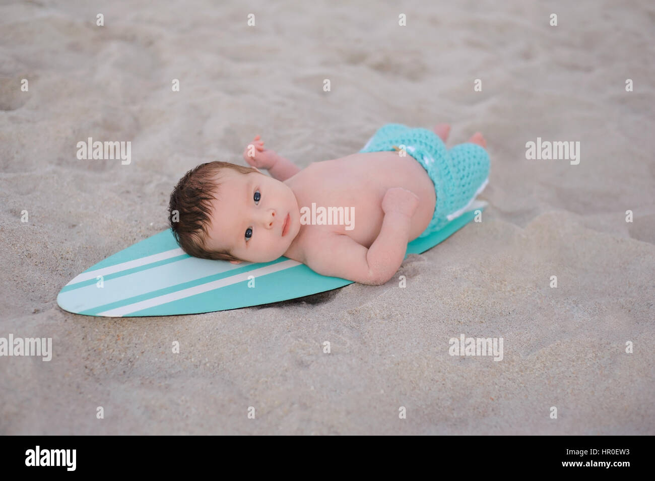 Two week old newborn baby boy lying on a tiny, turquoise blue and white surfboard. He is wearing aqua colored, board shorts and lying on a sandy beach Stock Photo
