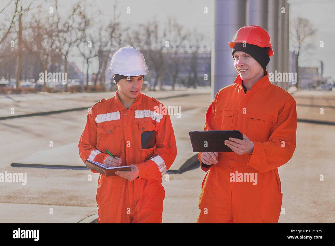 Cheerful young builders in orange uniform with the tablet and notepad. Horizontal outdoors shot. Stock Photo