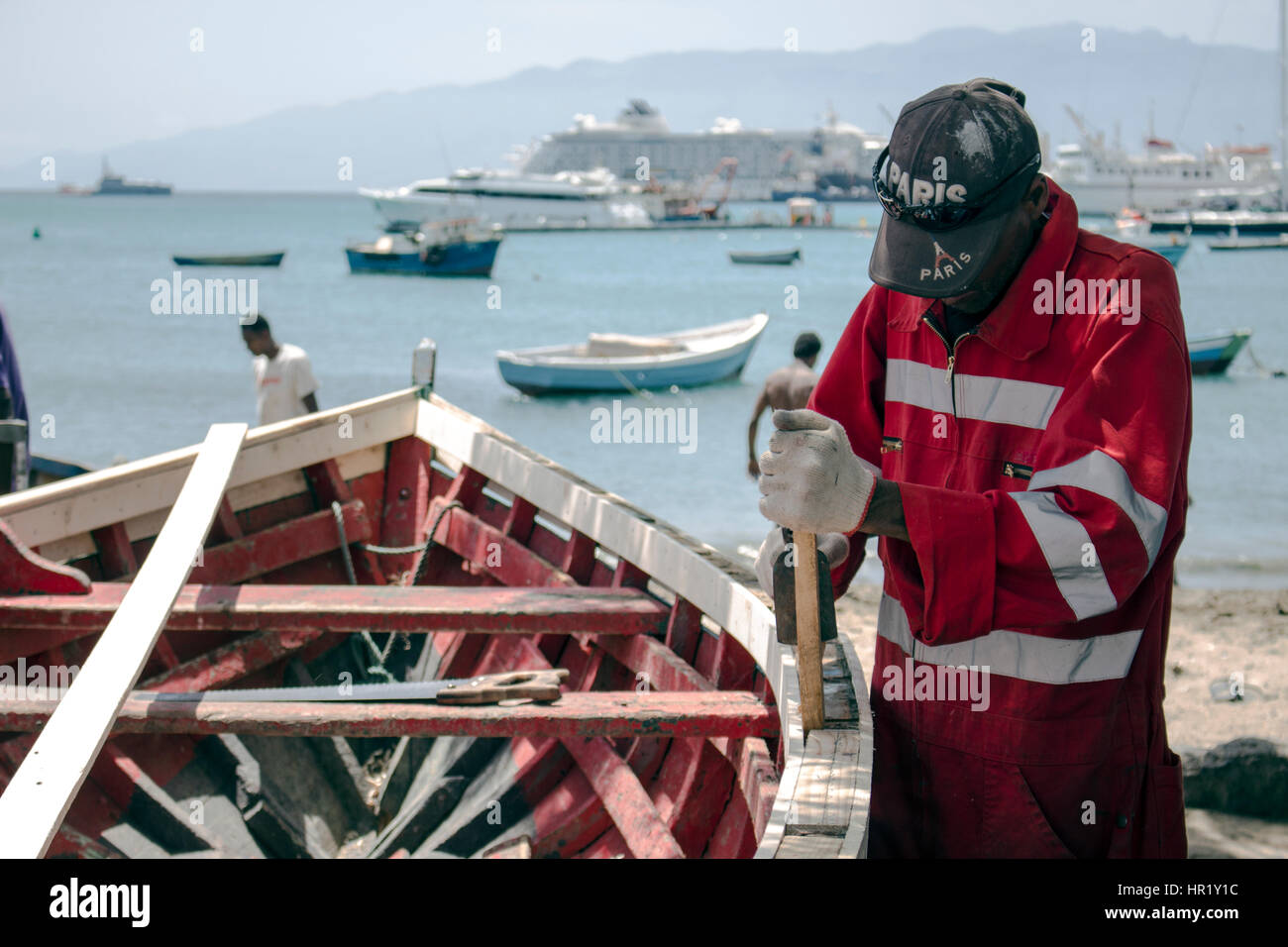 A worker carpenter is repairing a wooden boat wearing overall and gloves. Stock Photo