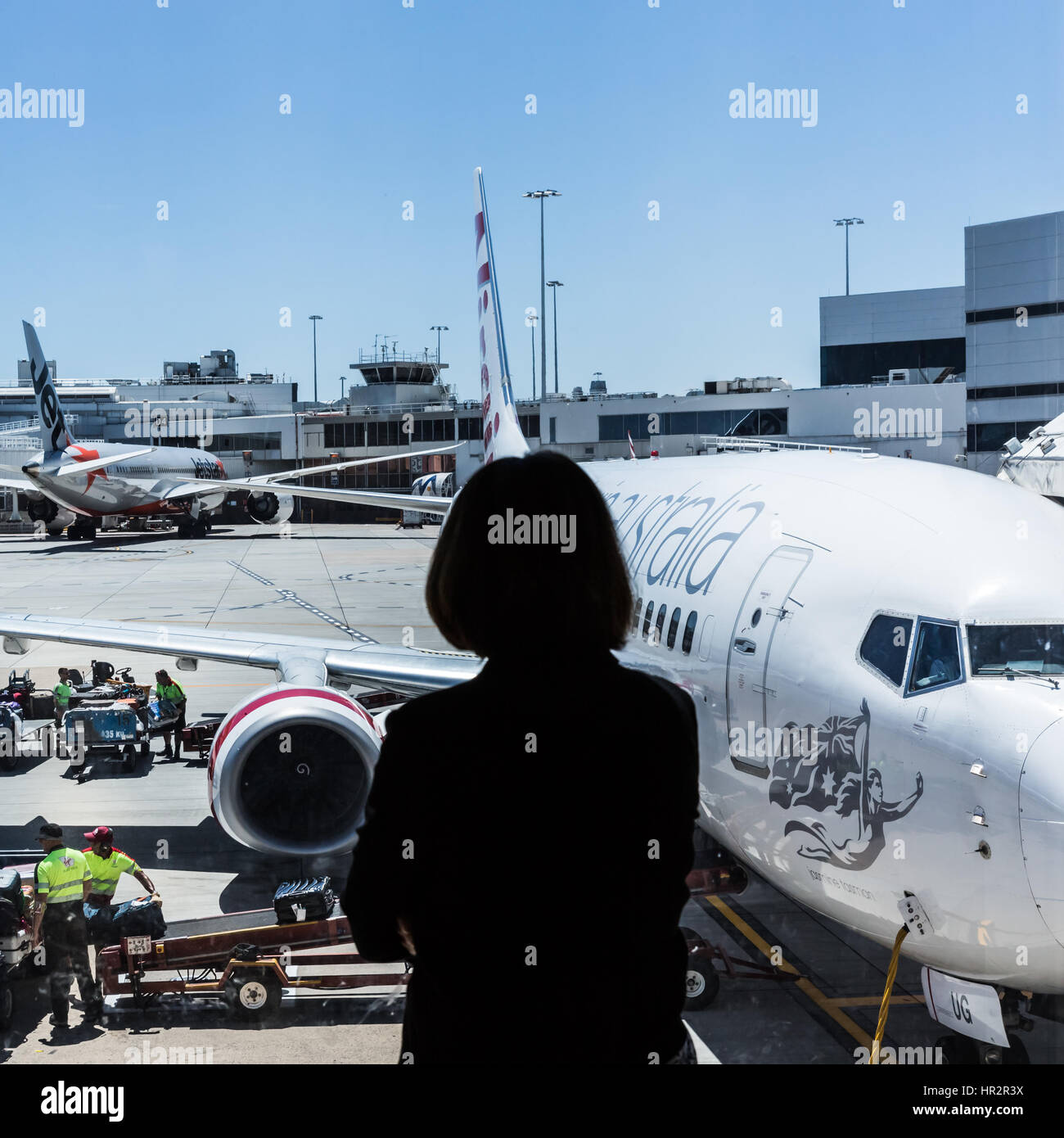 A woman in  silhouette wtaches baggage handlers loading an airplane at terminal. Stock Photo
