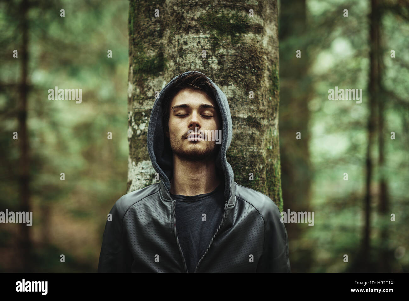 Young man in a forest leaning on a tree with eyes closed, individuality ...