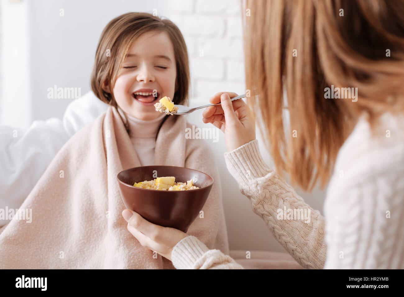 Everyday meal. Cheerful little girl sitting on the couch while her loving mother giving her breakfast Stock Photo