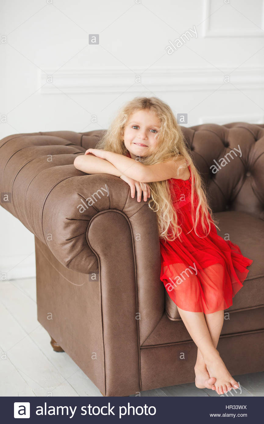 A Little Girl With Curly Blond Hair In A Red Dress Sitting On An