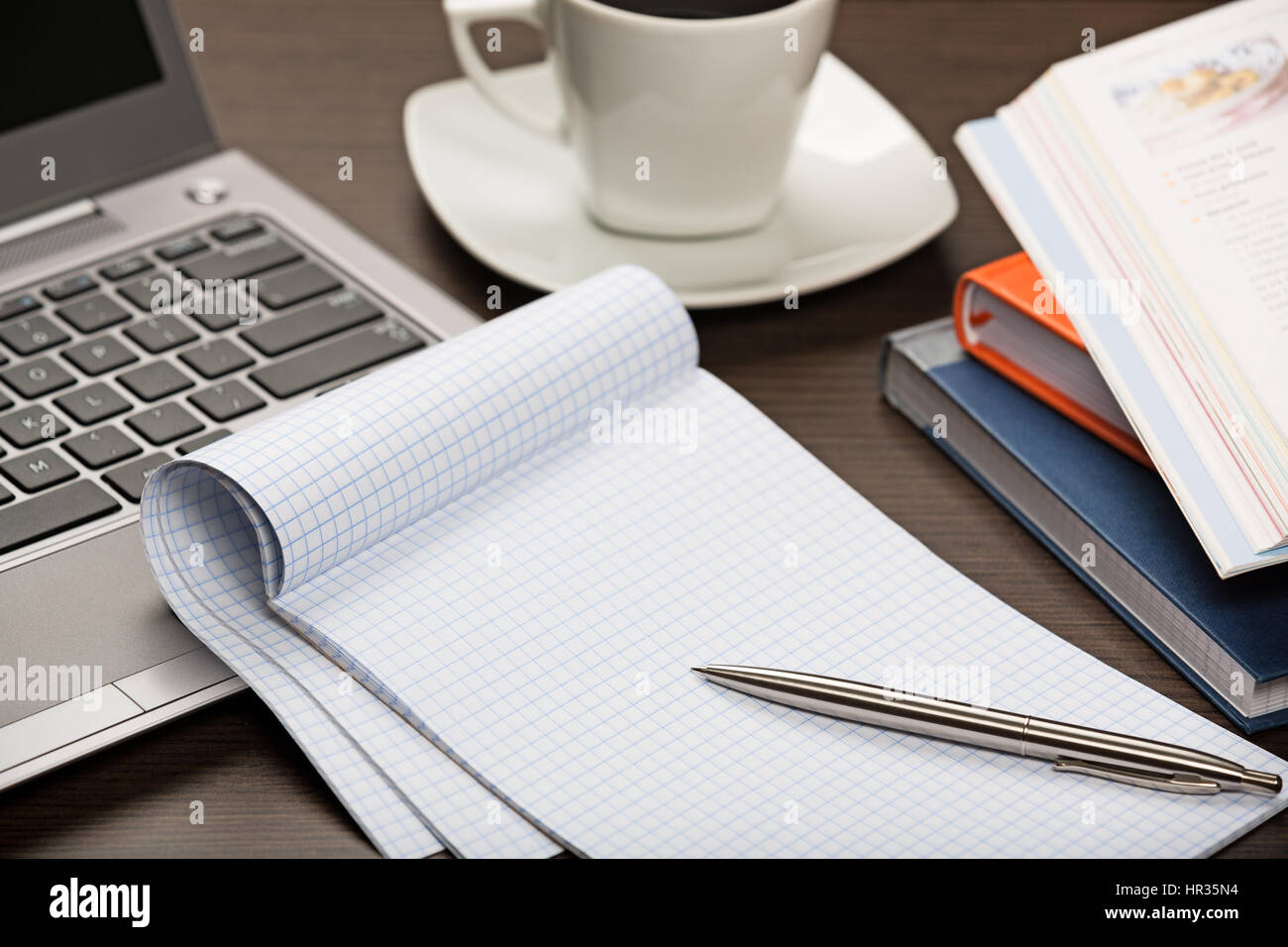 Notepad, pen, books, laptop and cup of coffee on dark wooden office