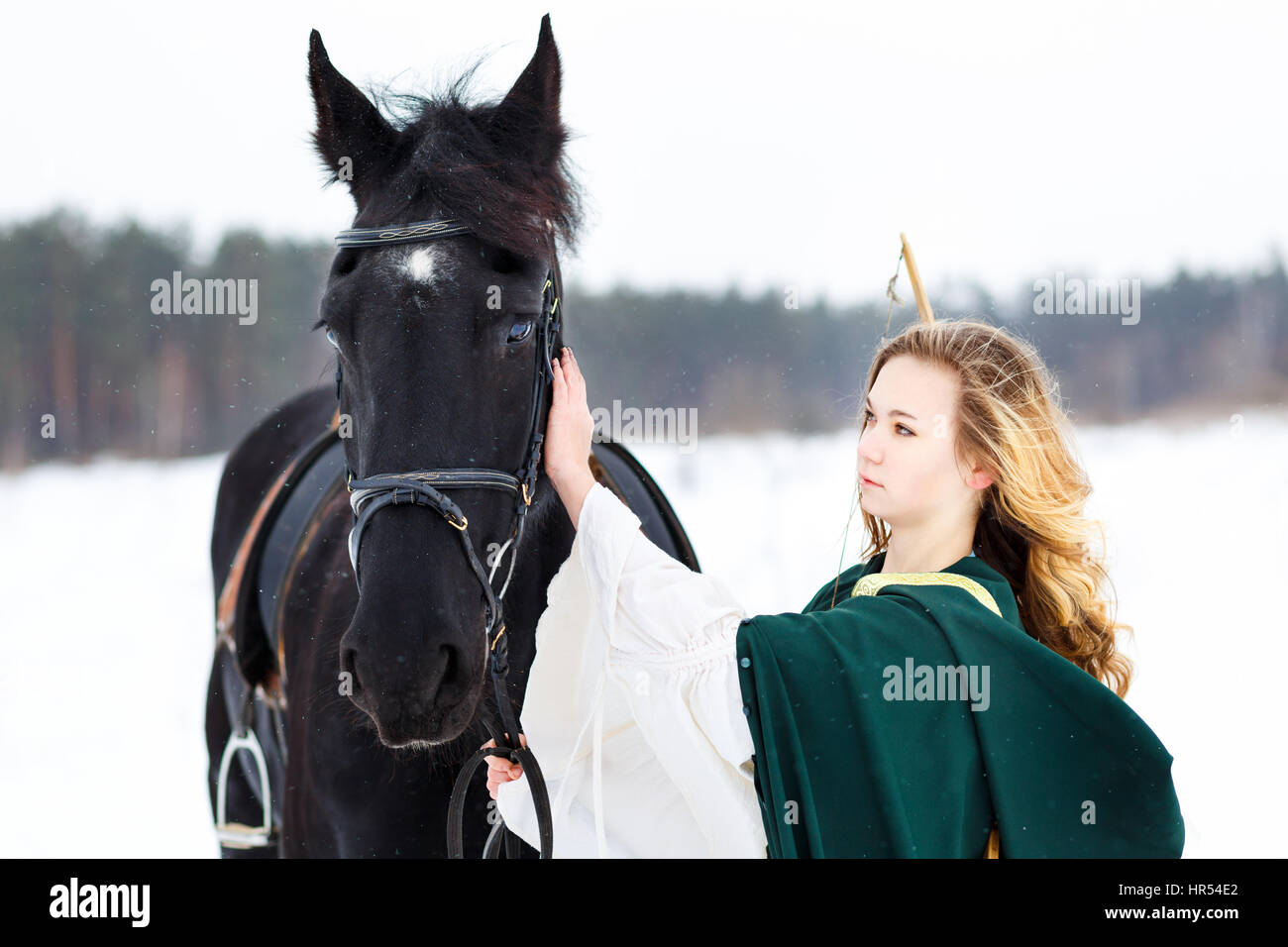 Young girl in white dress and green cape with black thoroughbred horse on winter field. Historical image Stock Photo