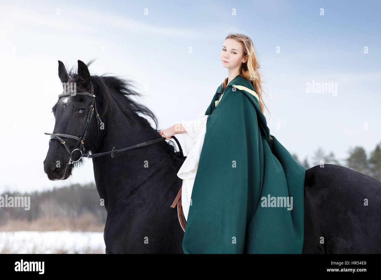 Young girl in white dress and green cape riding black thoroughbred horse in winter. Historical image Stock Photo