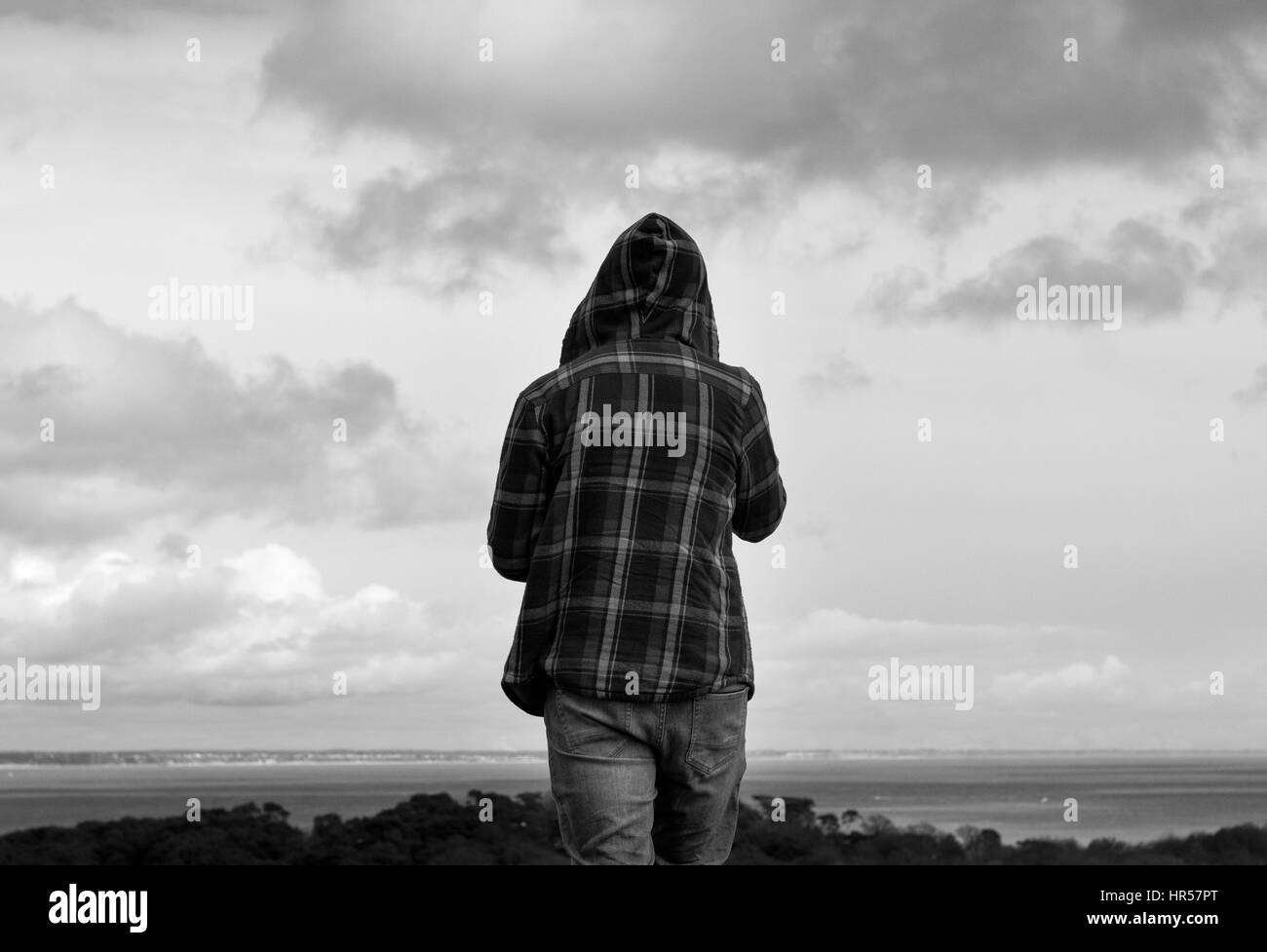 Black and white image of a hooded young man from behind looking over a landscape. Stock Photo