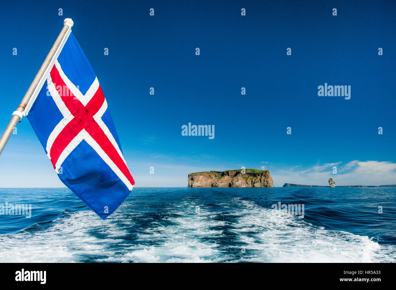 View from boat with Icelandic flag with Drangey Island in background over blue ocean. Stock Photo