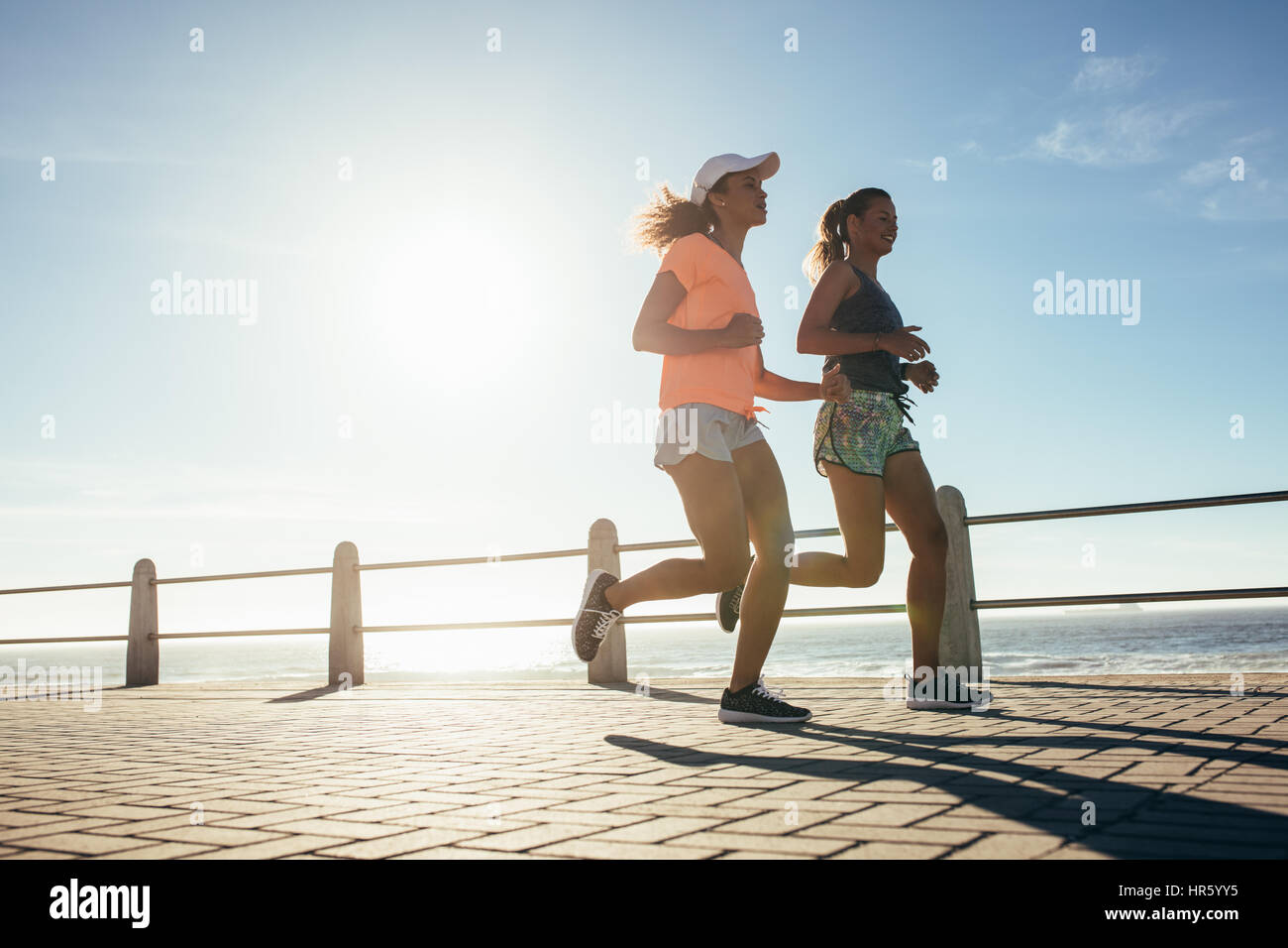 Low angle shot of two young women running on ocean front. Female runner working out on a sunny day. Stock Photo