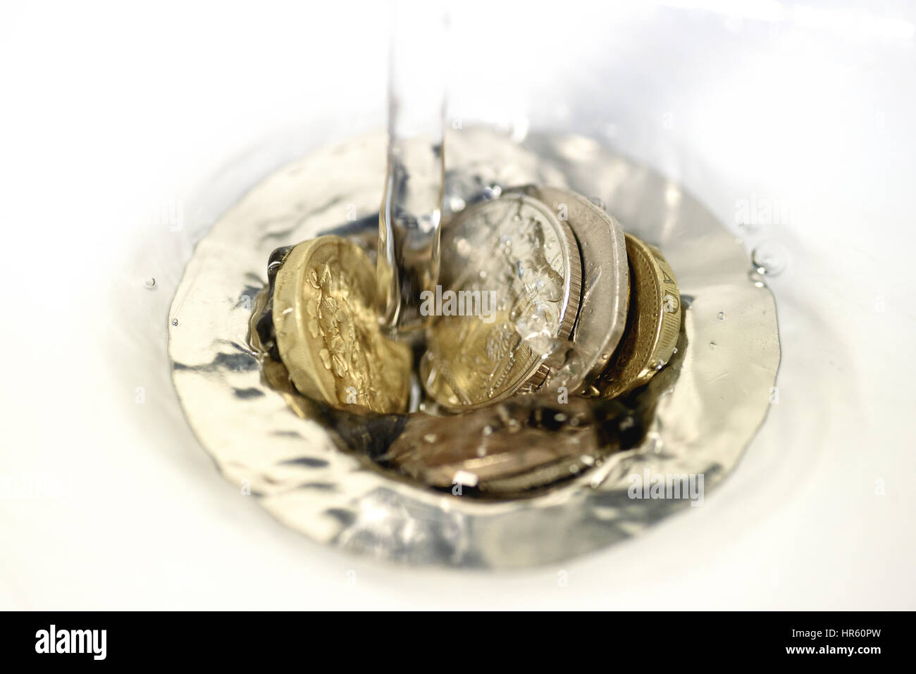 British money including a one pound coin stuck in a sink drain plughole as water flows over the coins. Stock Photo